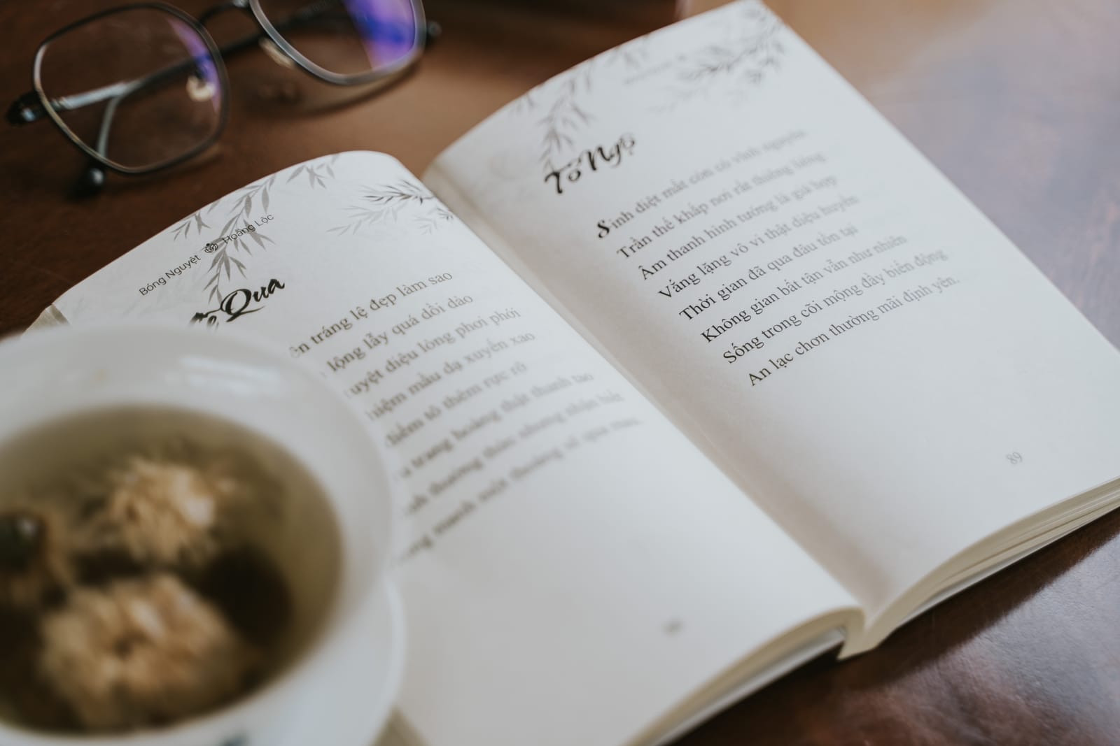 Cuốn thơ mở sẵn bên cặp kính và chén trà trên bàn gỗ.